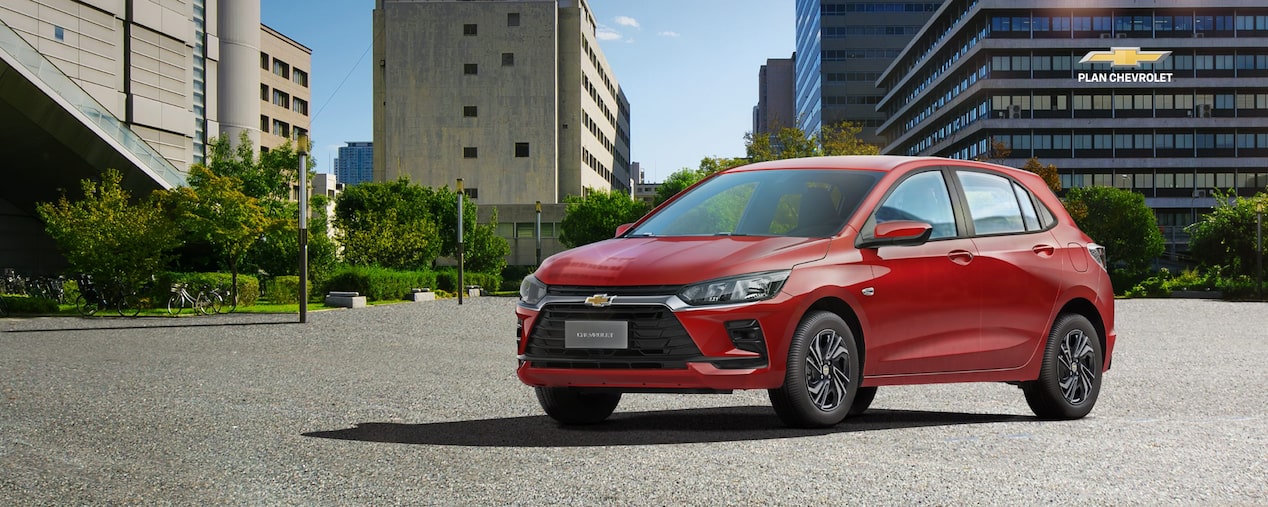 Chevrolet Onix rojo estacionado en un entorno urbano moderno, rodeado de edificios y áreas verdes. La imagen promociona financiación de hasta el 100% para un vehículo 0 km a través de Plan Chevrolet.