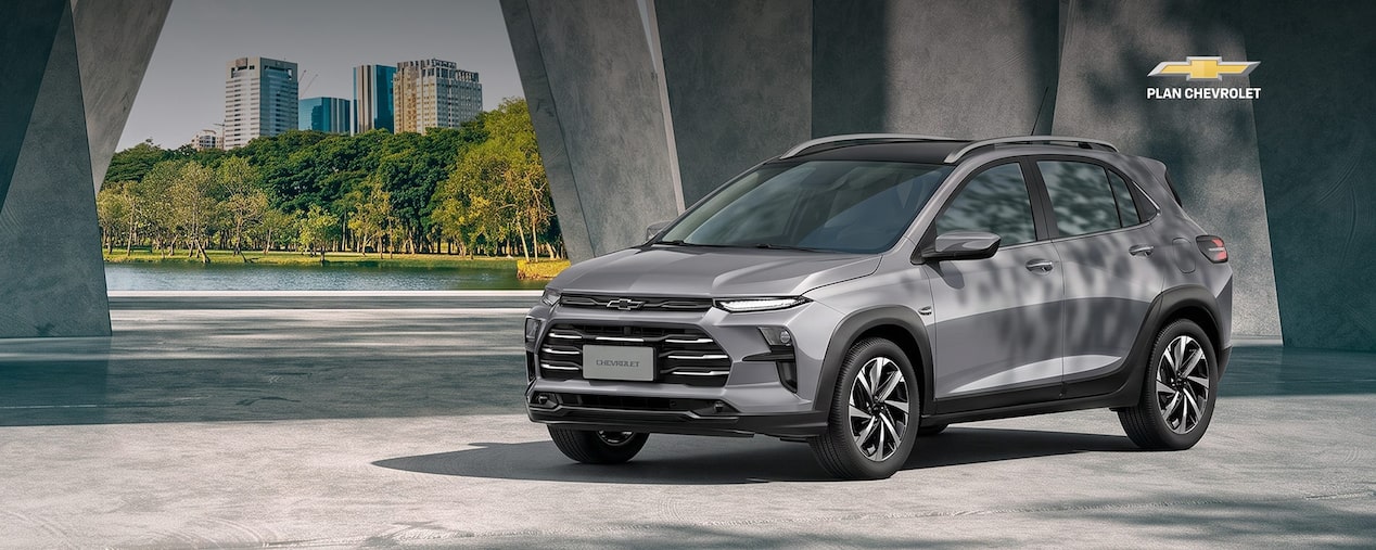 Chevrolet Sonic gris estacionado bajo una estructura de hormigón moderna, con un parque y edificios al fondo. La imagen promociona financiación de hasta el 100% en 84 cuotas con adjudicación pactada dentro de Plan Chevrolet.