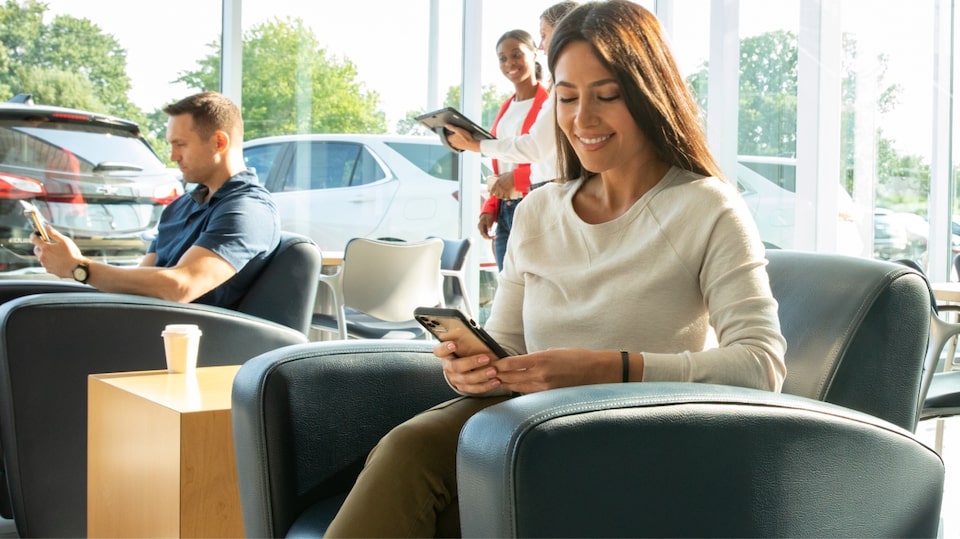 Persona usando un smartphone en una sala de espera moderna durante el día, con otras personas al fondo utilizando dispositivos, imagen que transmite la facilidad de gestionar y cobrar suscripciones digitales Chevrolet en línea dentro de la sección 'Cobrar Haber Neto'.
