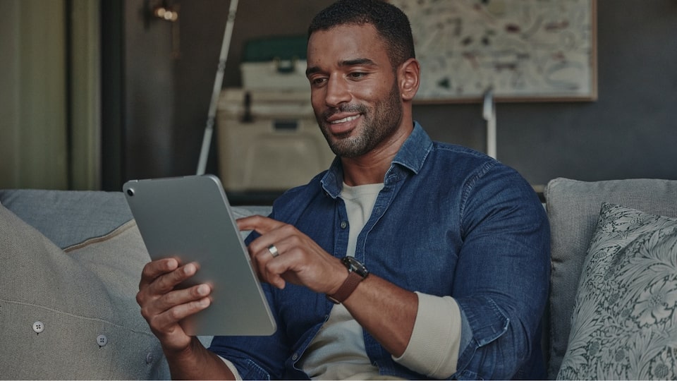 Persona sentada en un sofá usando una tablet, junto al texto 'Grupos participantes y modalidades de adjudicación', imagen que representa la consulta digital de listados y modalidades de adjudicación en la suscripción digital Chevrolet.