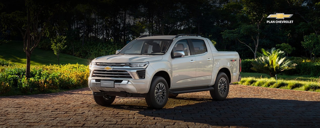 Chevrolet S10 blanca estacionada en un entorno residencial con áreas verdes y jardines al fondo. Imagen promocional de Plan Chevrolet con oferta de financiación de hasta 100% en 84 cuotas y adjudicación pactada.