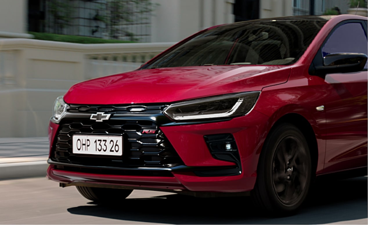 Vista exterior frontal en movimiento del nuevo auto Chevrolet Onix Hatchback 2027 color rojo circulando por la ciudad, acompañando la sección para explorar ofertas y condiciones comerciales en Argentina.