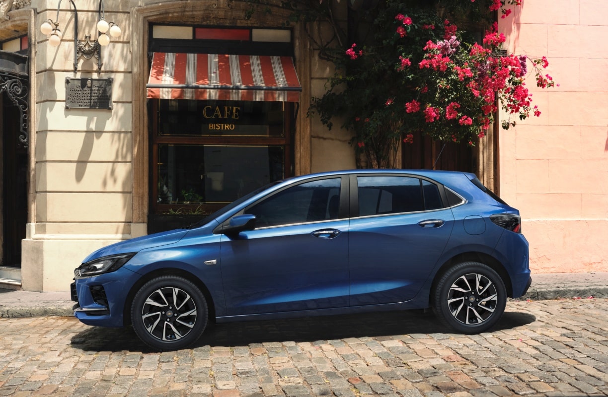 Vista exterior de perfil del nuevo auto Chevrolet Onix Hatchback 2027 color azul estacionado frente a un café, reflejando su agilidad, eficiencia y diseño ideal para la ciudad en Argentina.