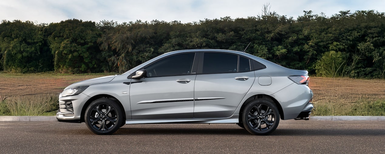 Imagen lateral de un Chevrolet Onix Plus, estacionado sobre pavimento al aire libre, con árboles y naturaleza de fondo. 