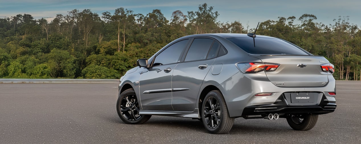 Vista trasera de un Chevrolet Onix Plus estacionado al aire libre, sobre pavimimento, con árboles y naturaleza de fondo. 
