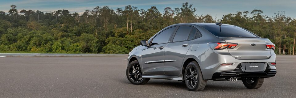 Vista trasera de un Chevrolet Onix Plus estacionado al aire libre, sobre pavimimento, con árboles y naturaleza de fondo. 