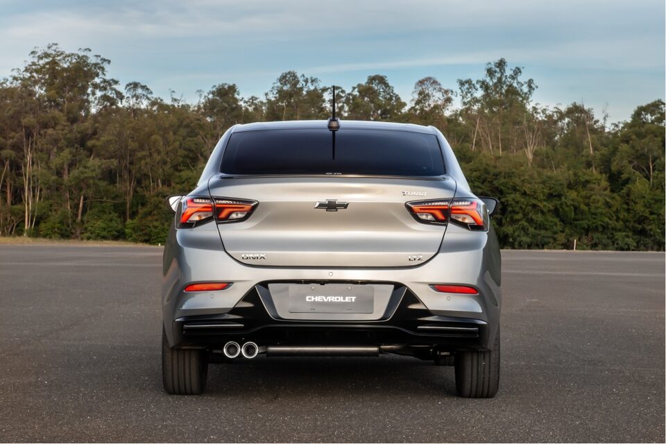Vista trasera del Chevrolet Onix Plus, estacionado al aire libre, sobre pavimimento, con árboles y naturaleza de fondo. 