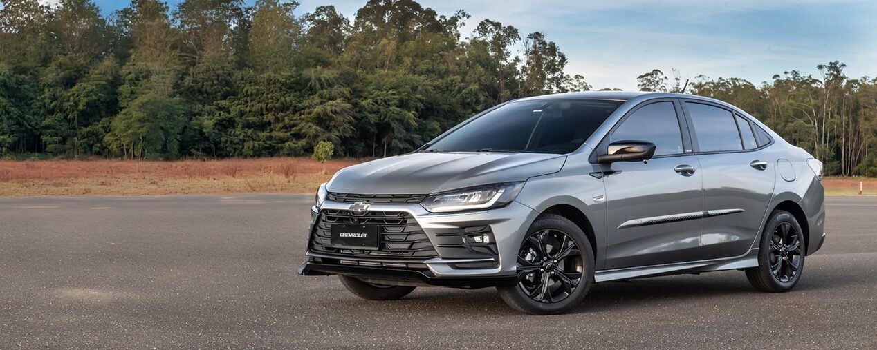 Imagen de un Chevrolet Onix Plus estacionado al aire libre, sobre pavimimento, con árboles y naturaleza de fondo. 