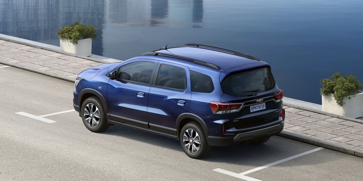 Vista trasera elevada en tres cuartos de la Chevrolet Spin 2027 azul con barras de techo estacionada junto al agua, destacando su motor 1.8L y diseño posterior con ópticas LED en Argentina.