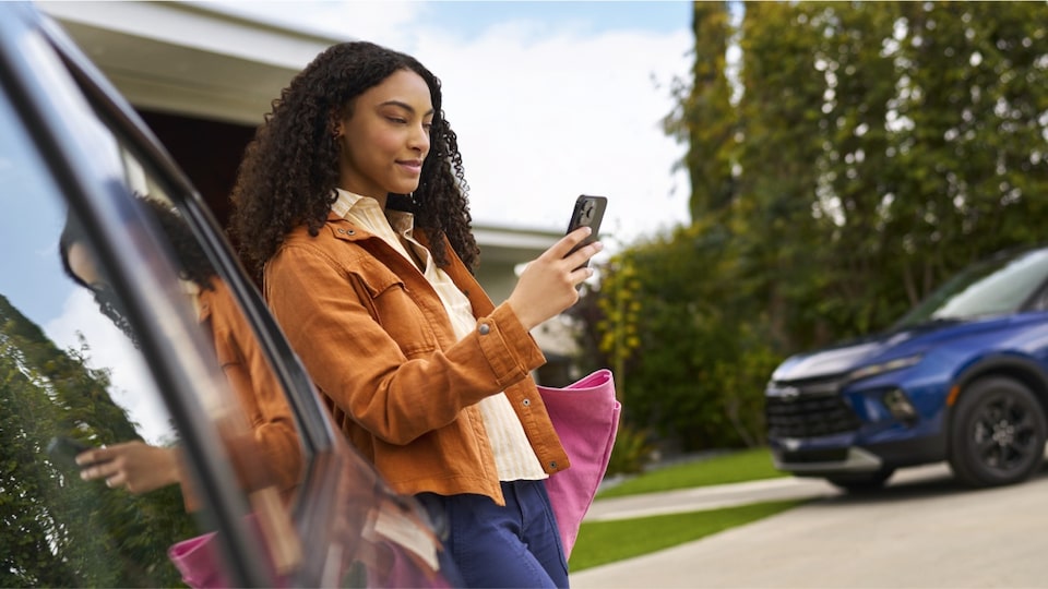 Imagen de una mujer usando su celular junto a su auto Chevrolet estacionado frente a una casa.