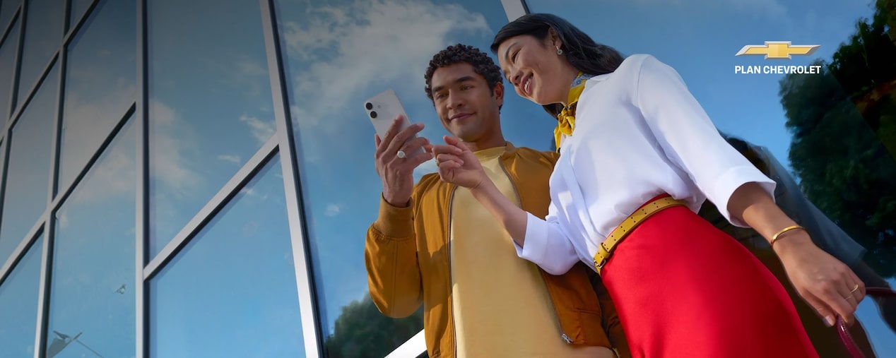 Dos personas revisan información de Plan Chevrolet desde un celular frente a un edificio corporativo.