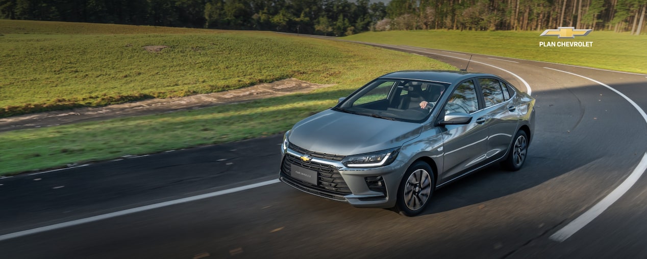 Chevrolet Onix Plus gris circulando por una carretera rodeada de áreas verdes. Imagen promocional de Plan Chevrolet con oferta de financiación de hasta 100% en 120 cuotas y seguro bonificado al 50% durante los primeros 3 meses.