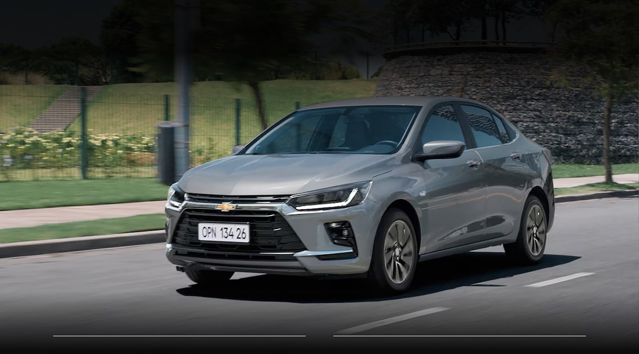 Chevrolet Onix Plus LT Turbo MT gris circulando por una avenida urbana rodeada de áreas verdes. Imagen promocional de Plan Chevrolet con información de financiación de hasta 80% en 120 cuotas y seguro bonificado al 50% durante los primeros 3 meses.