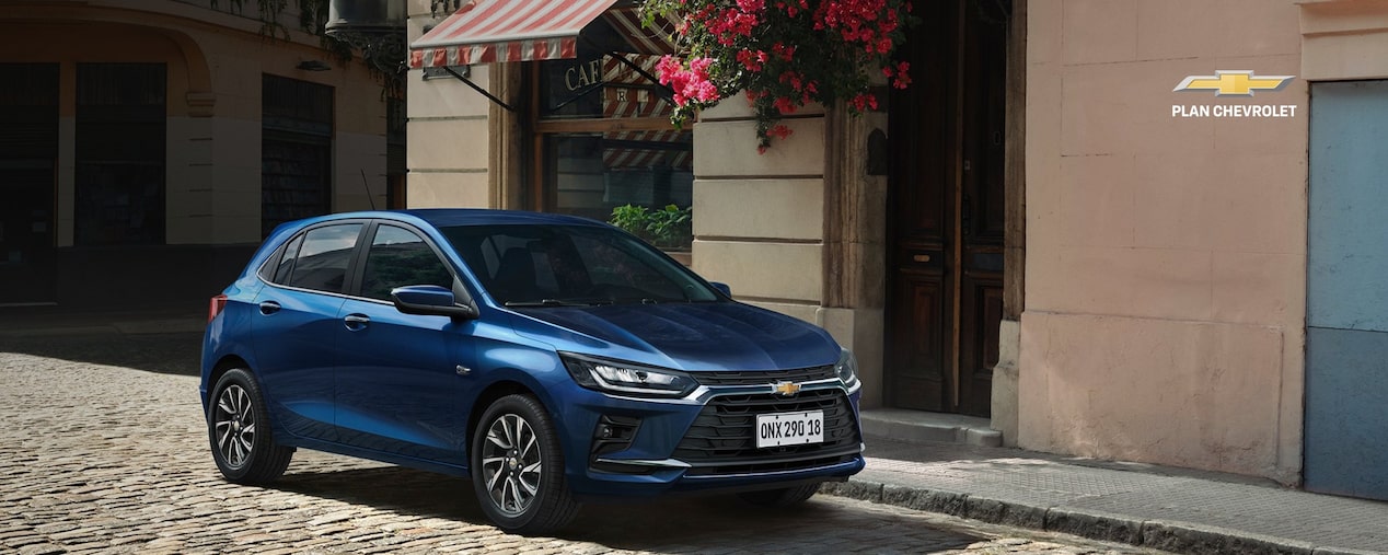 Chevrolet Onix azul estacionado en una calle adoquinada frente a edificios urbanos y comercios. Imagen promocional de Plan Chevrolet con oferta de financiación de hasta 80% en 120 cuotas y seguro bonificado al 50% durante los primeros 3 meses.