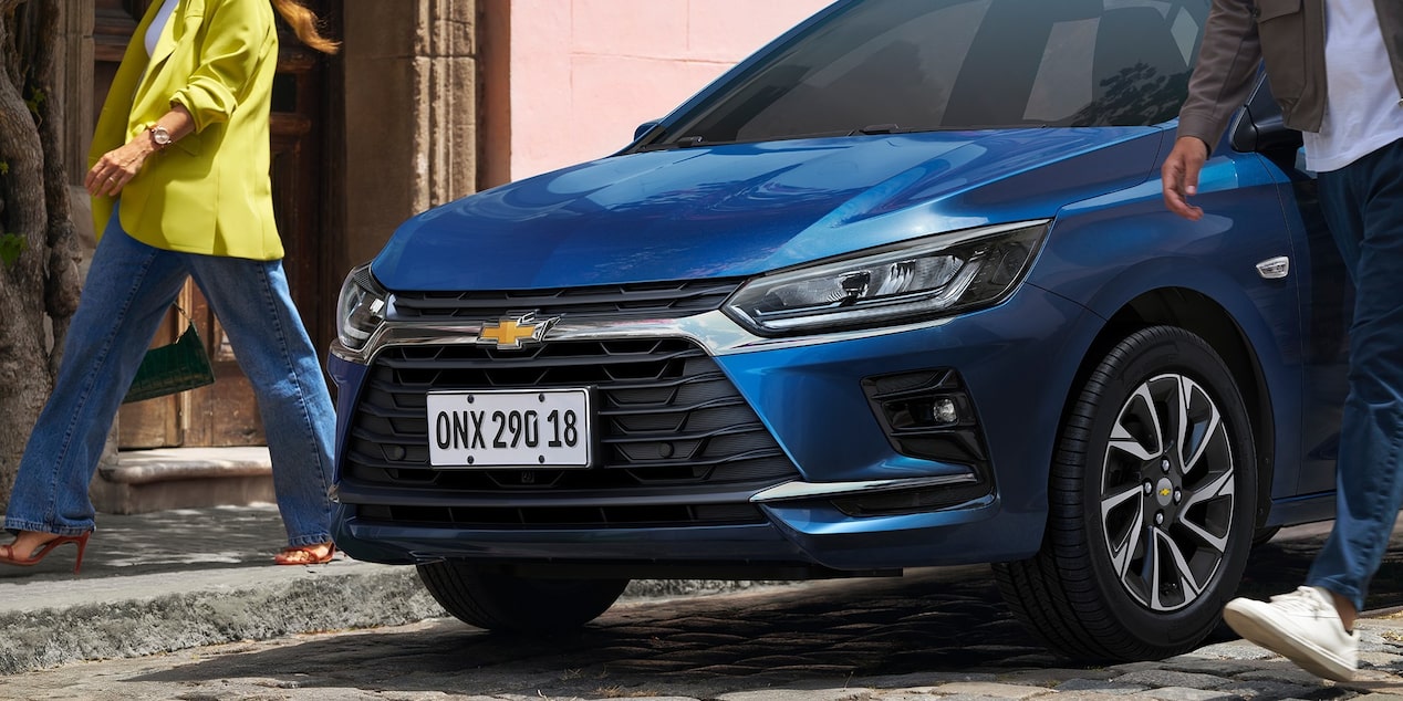 Detalle de la parrilla frontal con acabados cromados de Chevrolet Onix azul, estacionado en una calle urbana con personas caminando a su alrededor.