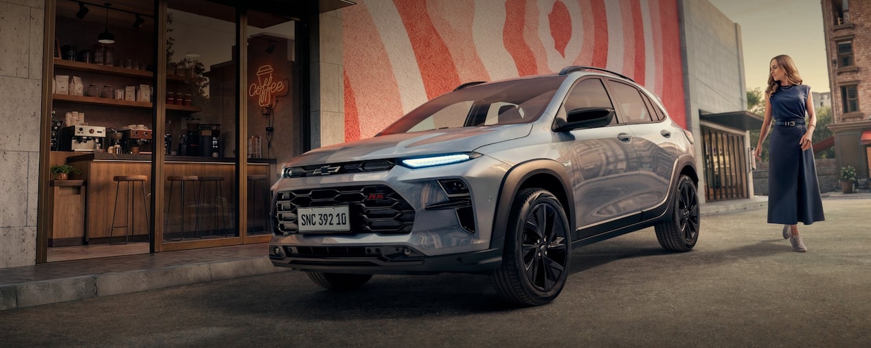 Un Chevrolet Sonic está estacionado frente a una cafetería. Una mujer camina junto al auto. Al fondo, se ve una pared decorada con obras de arte en tonos rojos. En la esquina superior izquierda, el texto “2027 - Sonic”. En la esquina inferior derecha, un botón: Reservá el tuyo.