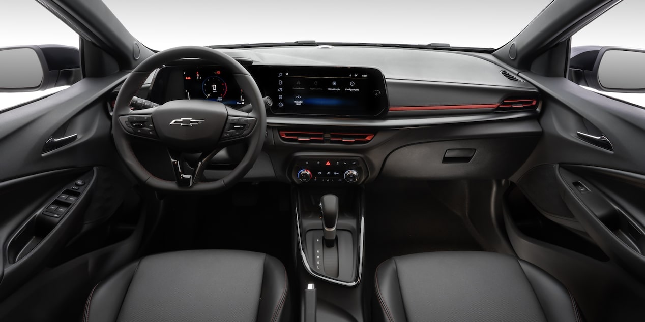 Vista interior y delantera del Chevrolet Sonic, mostrando volante, controles, pantalla central digital, aire acondicionado, palanca de cambios y guantera.