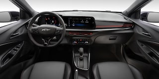 Vista interior y delantera del Chevrolet Sonic, mostrando volante, controles, pantalla central digital, aire acondicionado, palanca de cambios y guantera.