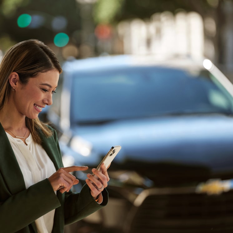Una mujer utilizando la app myChevrolet en su celular, con un auto Chevrolet detrás.