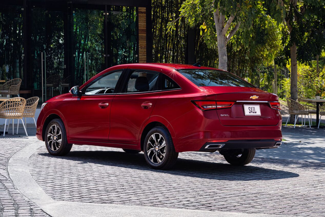 Vista trasera de la Chevrolet Sail Sedan estacionada, con árcoles, meses y sillas al fondo.