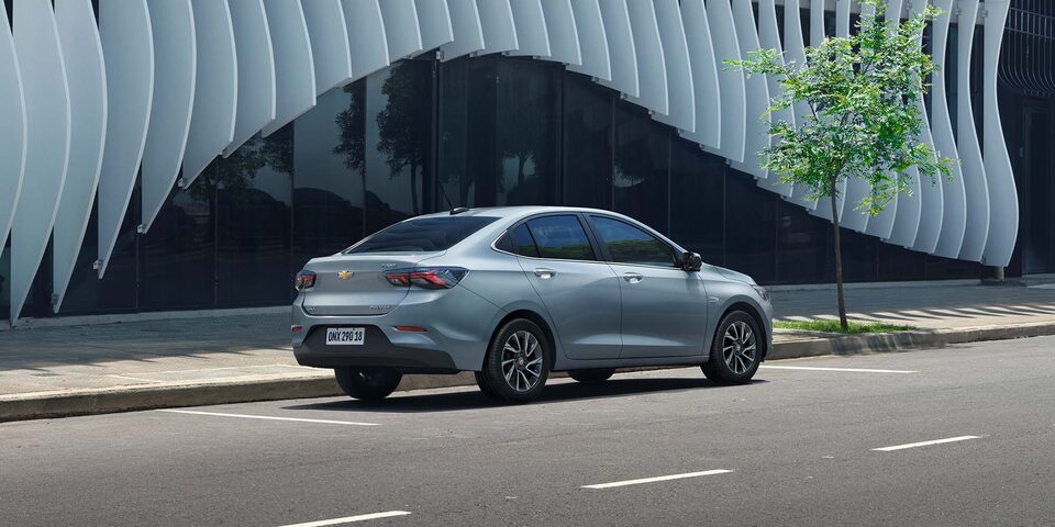 Chevrolet Onix Sedán estacionado en paralelo sobre una calle urbana, destacando el uso del asistente de parqueo semi-automático y su diseño moderno visto desde la parte trasera.