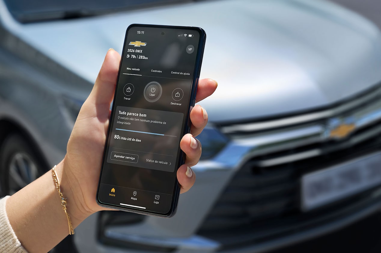 Persona sosteniendo un smartphone con la aplicación de myChevrolet conectada al Onix Sedán, mostrando en pantalla funciones de control remoto y diagnóstico del vehículo, con el auto visible al fondo.