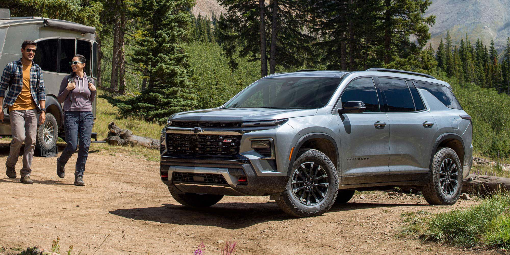 Un hombre y una mujer caminan sonrientes al lado de una Chevrolet Traverse Z71 estacionada en la base de una montaña, con pinos y montes al fondo. 