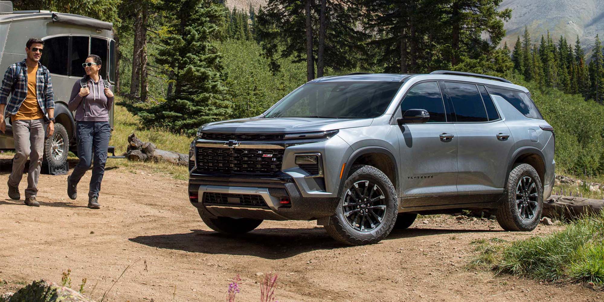Un hombre y una mujer caminan sonrientes al lado de una Chevrolet Traverse Z71 estacionada en la base de una montaña, con pinos y montes al fondo. 